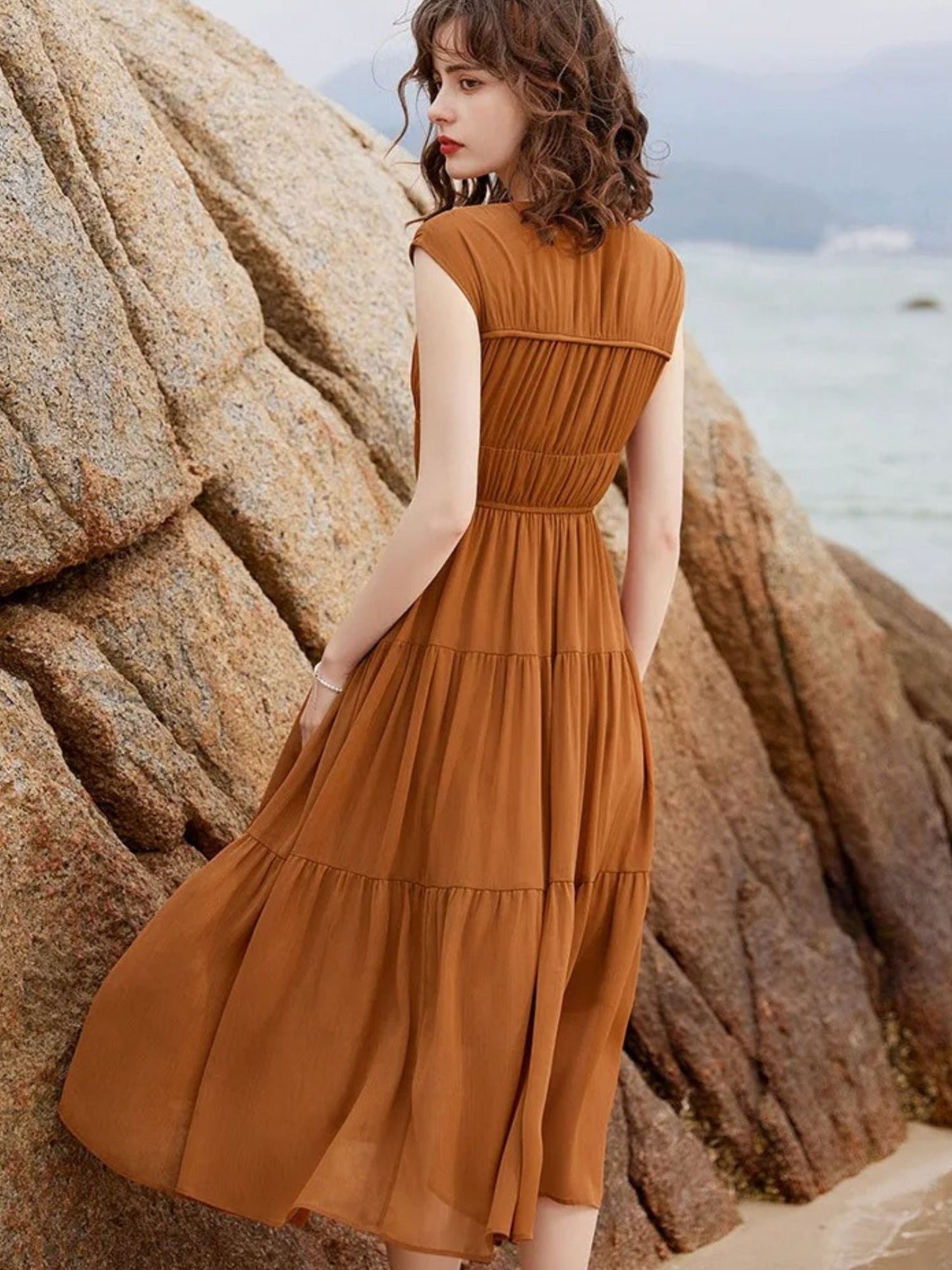 Rückansicht einer Frau im braunen ärmellosen Stufen-Maxikleid vor Felsen.