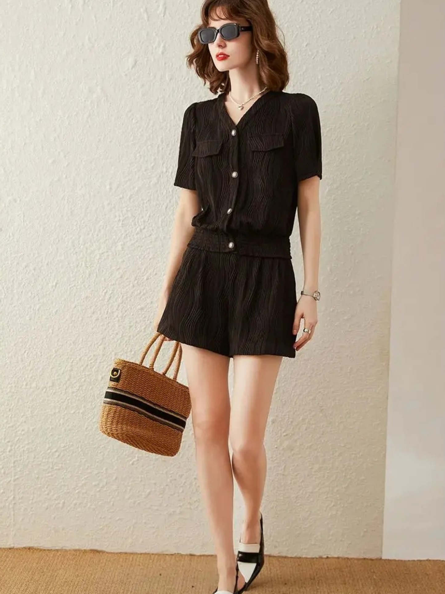 Frau posiert im schwarzen Jumpsuit mit Gürtel und Korbtasche.