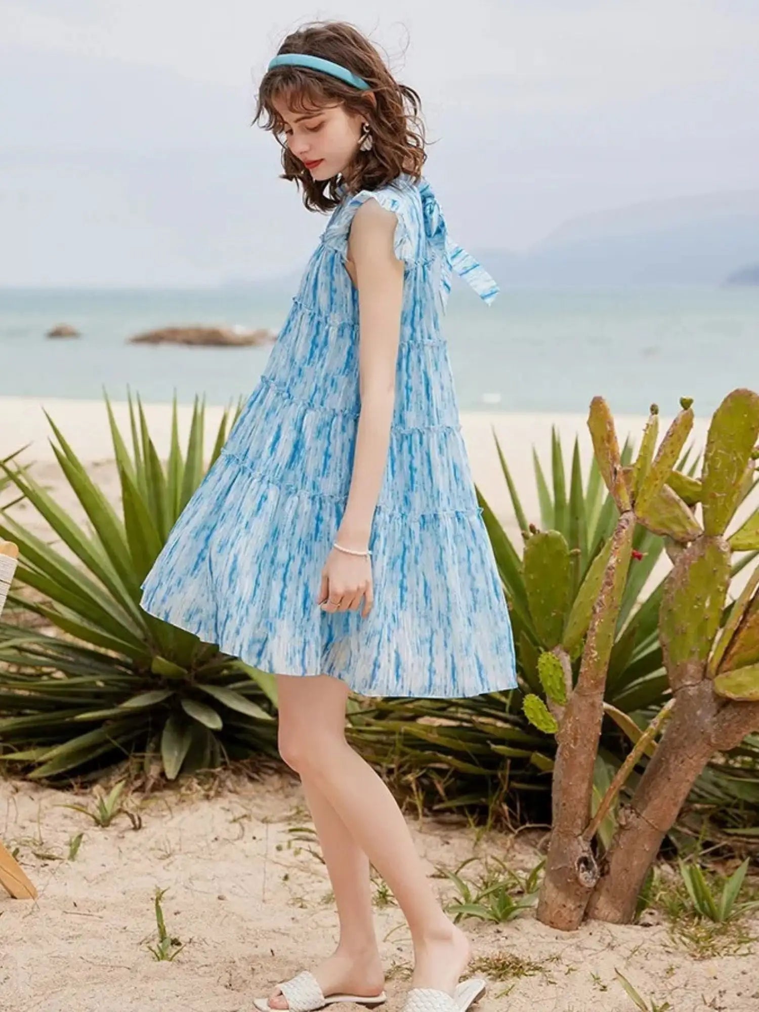 Frau geht am Strand im blauen Rüschenkleid mit Korbtasche.