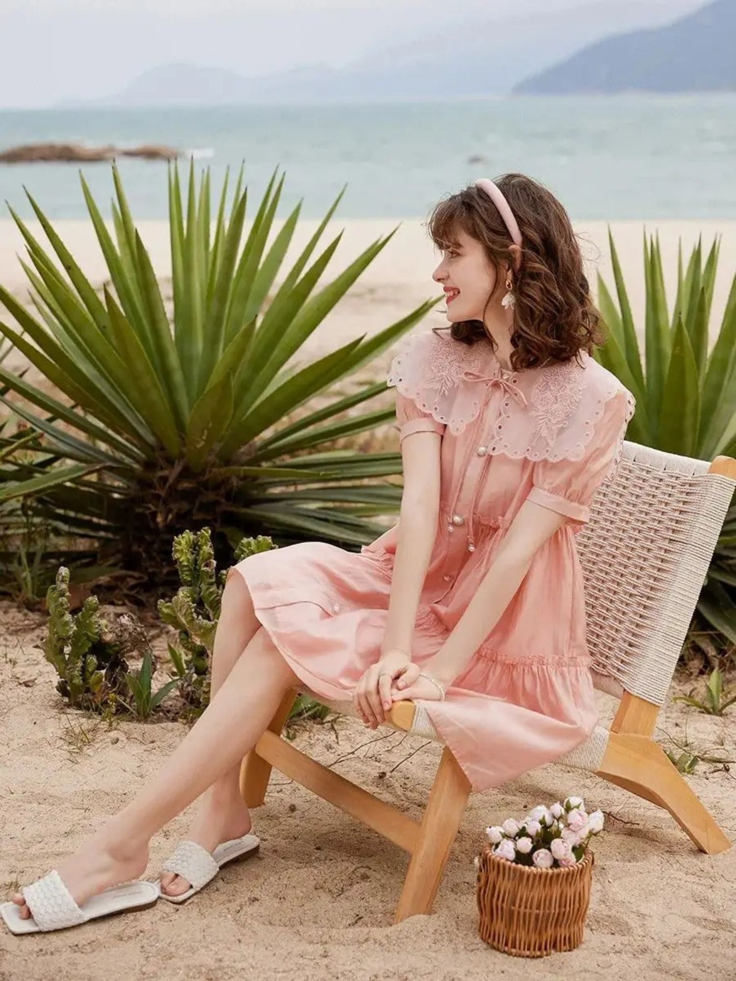 Frau im rosa Spitzenkleid sitzt am Strand mit Basttasche.